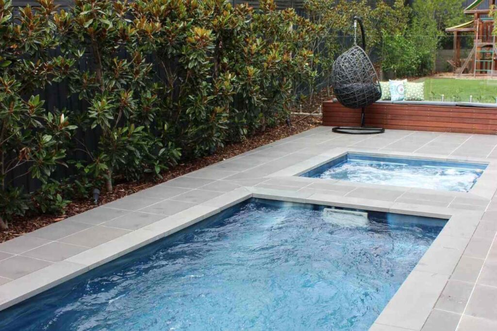 Custom-built plunge pool with adjacent heated spa section, surrounded by grey tile paving, privacy hedges, and a modern backyard seating area featuring a hanging egg chair and timber bench.