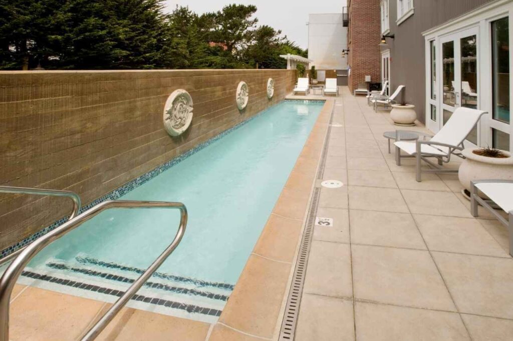 Narrow rectangular lap pool with light blue water, tiled edges, and stainless steel handrails, bordered by a decorative privacy wall and surrounded by poolside lounge chairs and planters on a tiled patio.