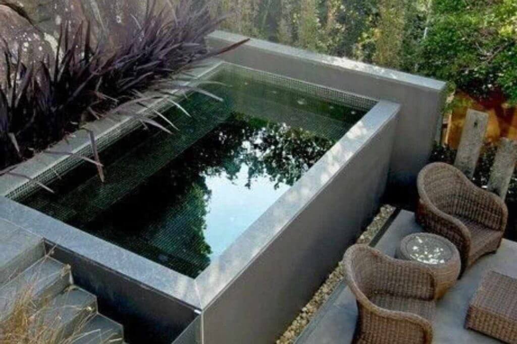 Sleek rectangular above-ground plunge pool with dark mosaic tiles, elevated concrete surround, and adjacent outdoor seating area featuring rattan chairs and a small table in a lush, private garden setting in Newcastle.