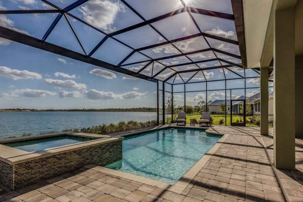 Enclosed backyard swimming pool and spa area with screened roof structure, overlooking a scenic lake, featuring lounge chairs, paver flooring, and a tranquil residential waterfront view.