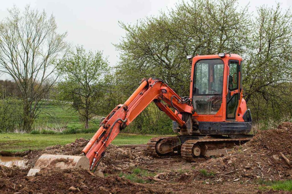 Orange excavator digging soil on a grassy block with surrounding trees, preparing the ground for plunge pool installation in a rural or suburban setting.