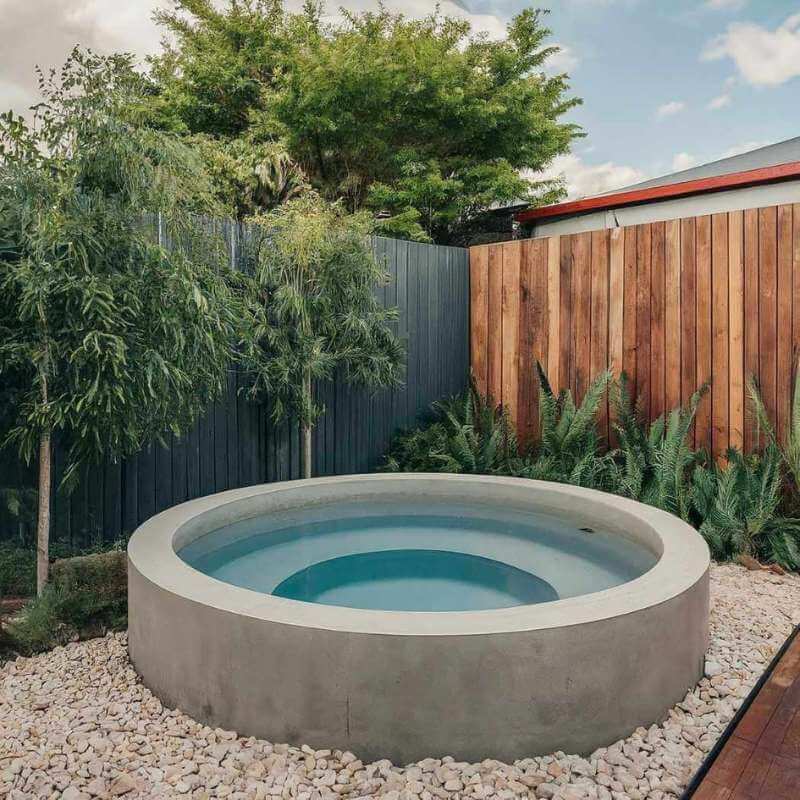 Modern above ground round precast concrete plunge pool surrounded by white pebbles and lush greenery, set in a private backyard with timber and metal fencing for a natural, spa-like retreat.