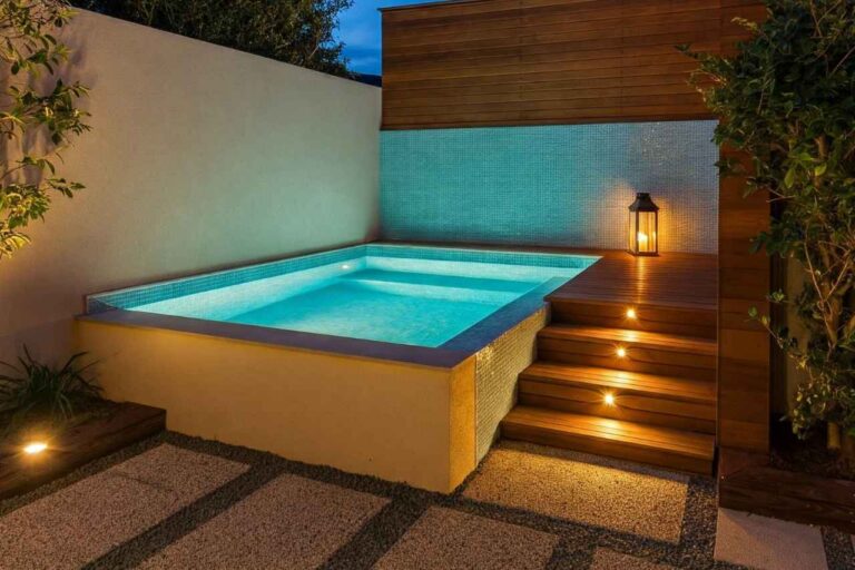 Modern above-ground plunge pool with soft blue underwater lighting, timber decking and steps, built-in wall tiles, and ambient lantern, set in a small landscaped backyard.