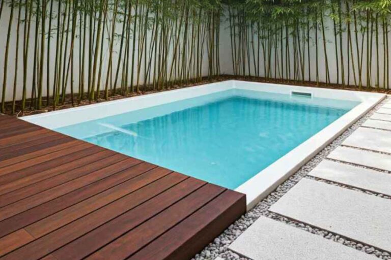 Modern inground plunge pool with sleek white edging, timber decking, and minimalist stepping stones, surrounded by a privacy wall lined with tall bamboo plants in a contemporary backyard setting.