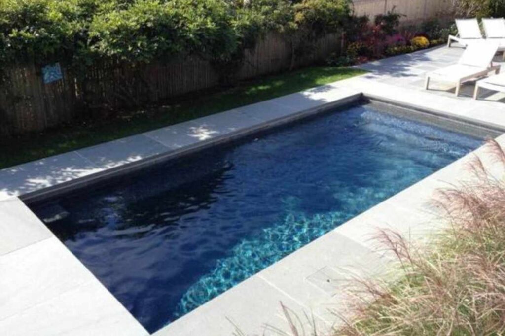Modern rectangular inground plunge pool with dark blue tiles, surrounded by light grey stone pavers, adjacent to a green lawn and white poolside loungers in a landscaped Newcastle backyard.
