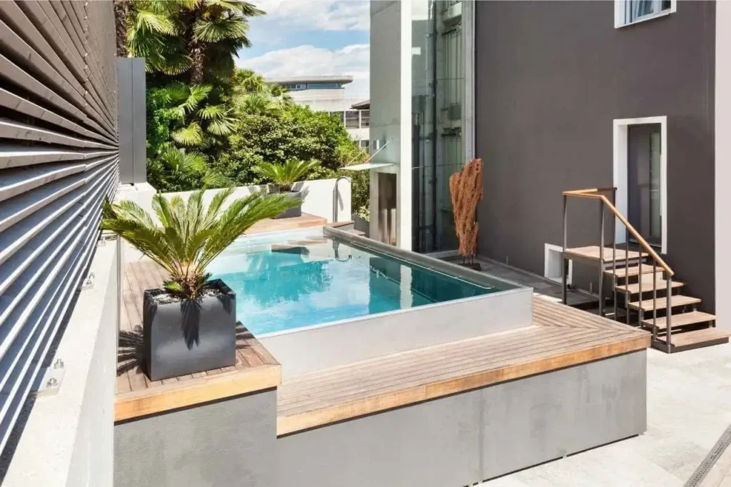 Modern semi-inground plunge pool with a sleek concrete exterior, timber decking, and glass fencing, set beside a contemporary grey building with tropical plants and outdoor stairs.