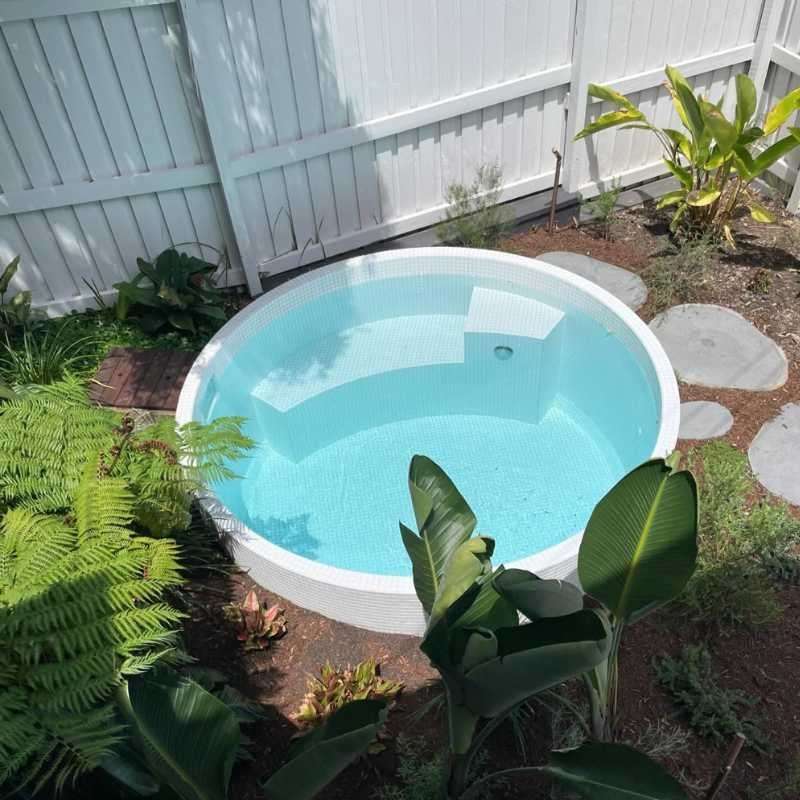 Round above ground plunge pool with light blue tiled interior surrounded by tropical plants and white fence in a compact backyard in Newcastle NSW