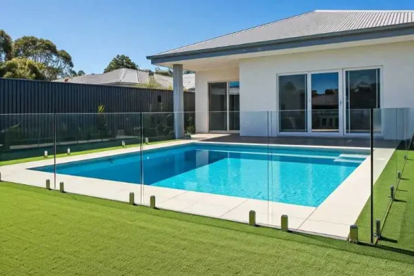 Contemporary backyard with a precast plunge pool surrounded by artificial turf and glass fencing, adjacent to a modern white home with large windows.
